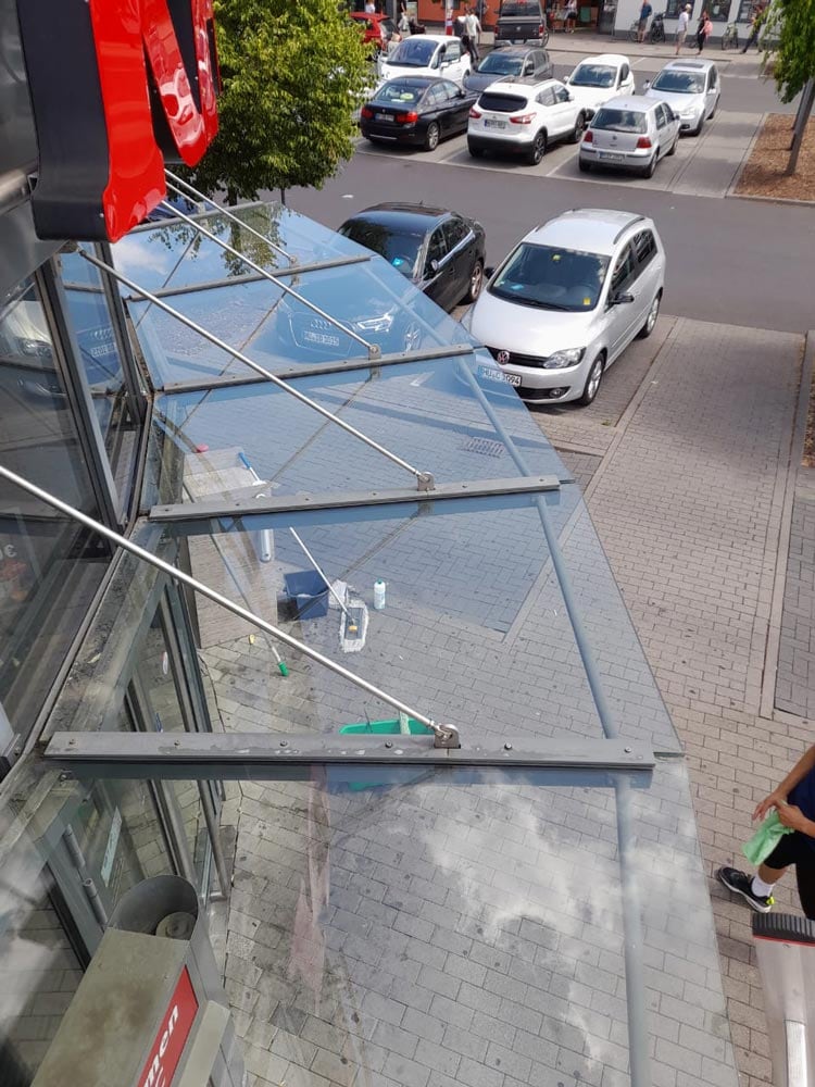 Eine Person reinigt Fenster eines Gebäudes. Autos sind im Hinter- und Vordergrund sichtbar.